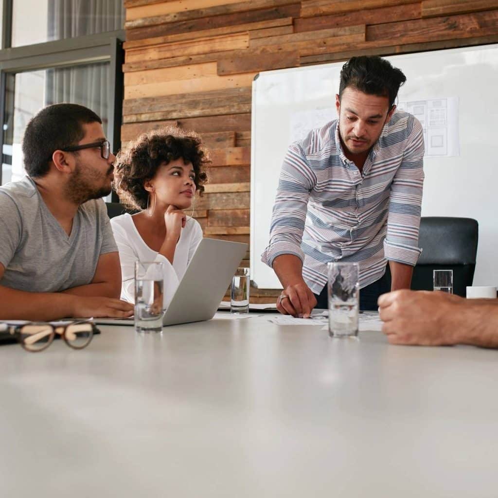 A man in a striped shirt conducts a business meeting at a conference table while three others listen.