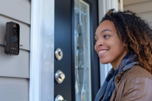 A smiling woman stands outside a front door equipped with a smart video doorbell and electronic smart lock system.