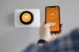 Person using a smartphone app to control a smart thermostat set to 77 degrees on a wall.