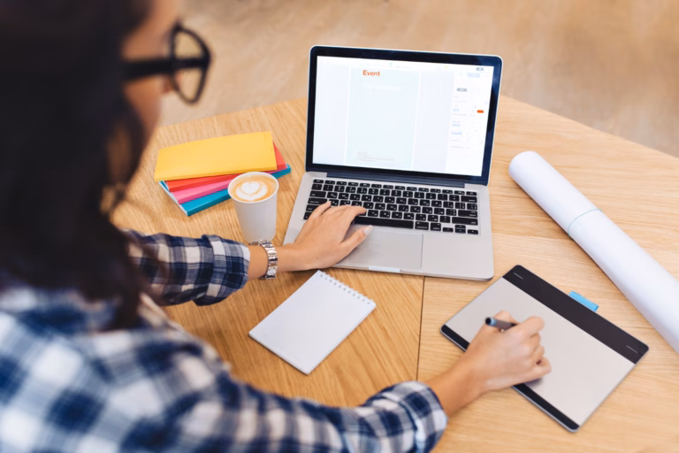 Small business owner working at a laptop with a notebook, coffee, and colorful folders on a wooden desk.