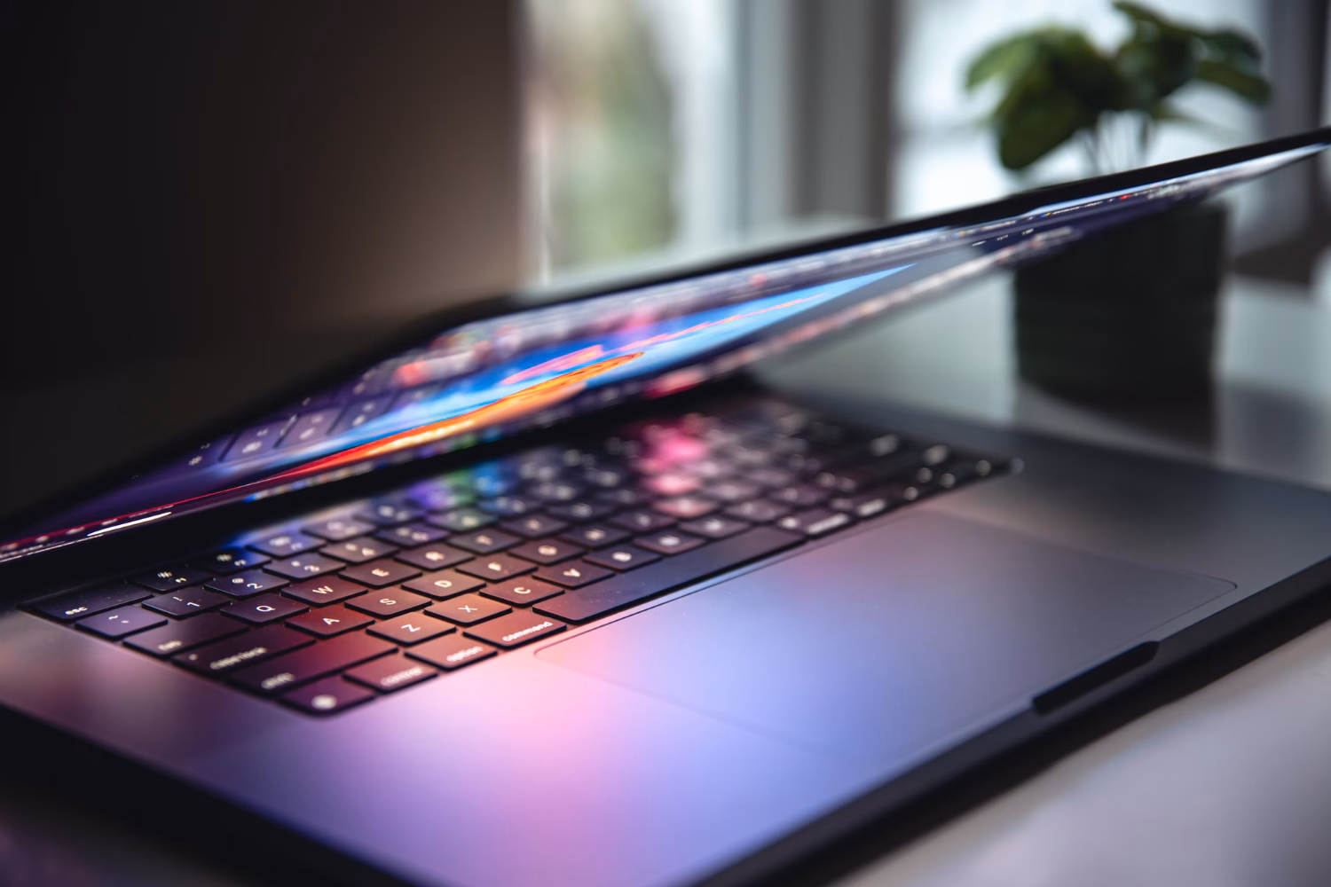 Close-up of a modern laptop with a partially closed screen, reflecting colorful light on the keyboard in a home office setting