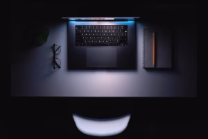 Overhead view of a sleek laptop on a dimly lit desk with glasses, a spiral notebook, and pencil — minimalist developer workspace at night.