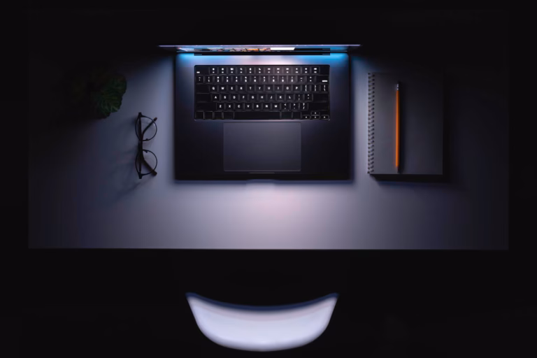 Overhead view of a sleek laptop on a dimly lit desk with glasses, a spiral notebook, and pencil — minimalist developer workspace at night.