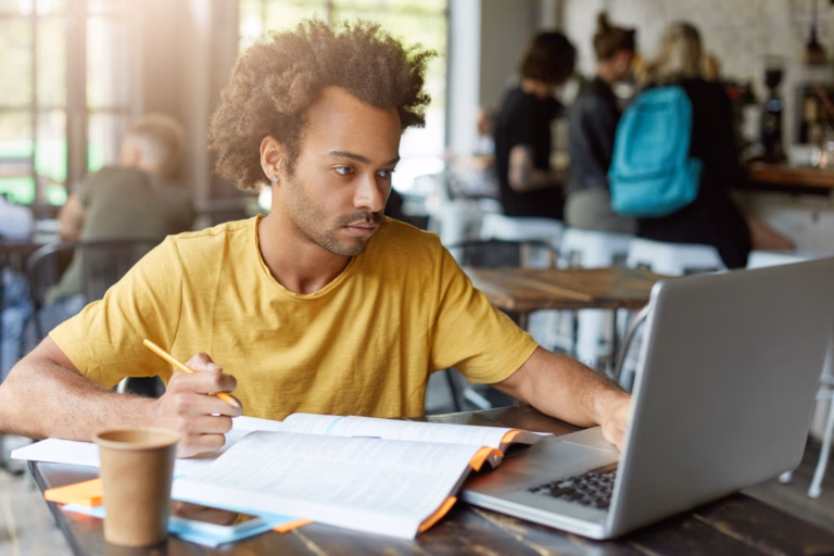 College student studying at a café with a laptop, textbook, and coffee — focused on schoolwork in a casual remote study environment