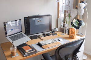 A real home office desk with a 24-inch monitor, laptop on a stand, mechanical keyboard, wireless mouse, desk lamp, small fan, and organized tech accessories