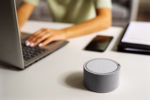 Smart speaker on a desk beside a laptop and smartphone, suggesting a home office setup.